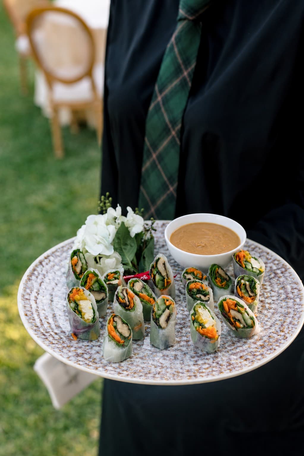 A plate of fresh vegetable spring rolls with a dipping sauce, held by a person wearing a black shirt and a green plaid tie.